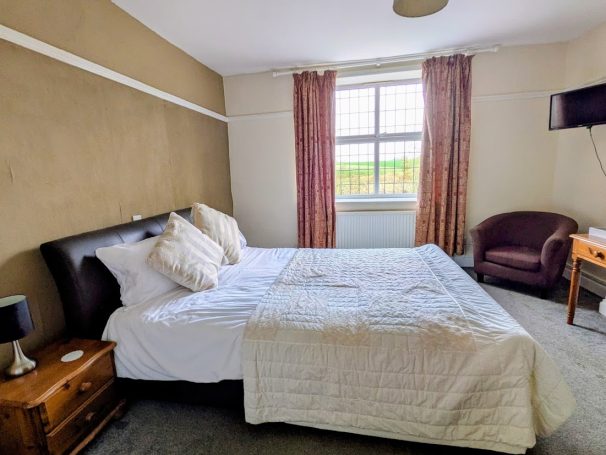 Double bed with leather headbord, arm chair and other wood furniture in large room with view of countryside out of the window.